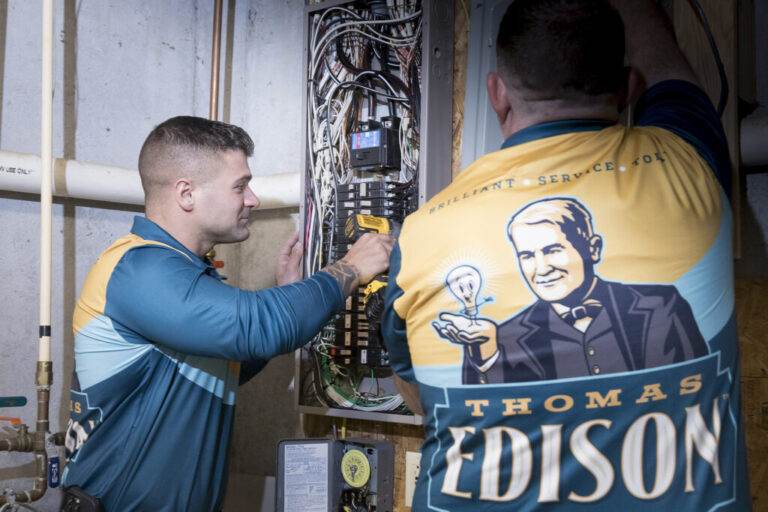 Two Thomas Edison Electric electricians testing circuits at an open residential electrical panel.