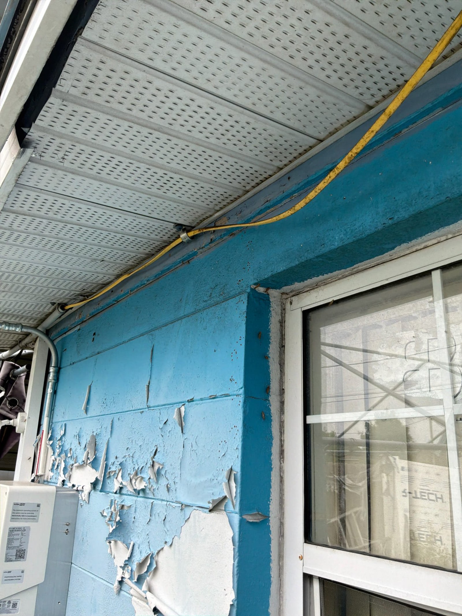 Exterior wall showing deteriorated painted surface with exposed yellow service conductor and corroded conduit.